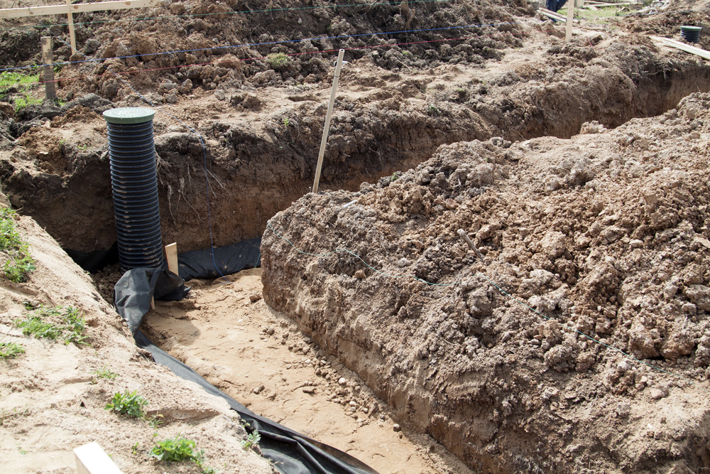 A construction site showing deep trenches in the soil, with exposed dirt and a black drainage pipe installed vertically near the trench wall.