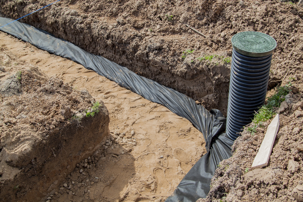 A trench lined with black fabric and a corrugated drainage pipe with a green cover installed vertically at the intersection of the trench on a construction site.