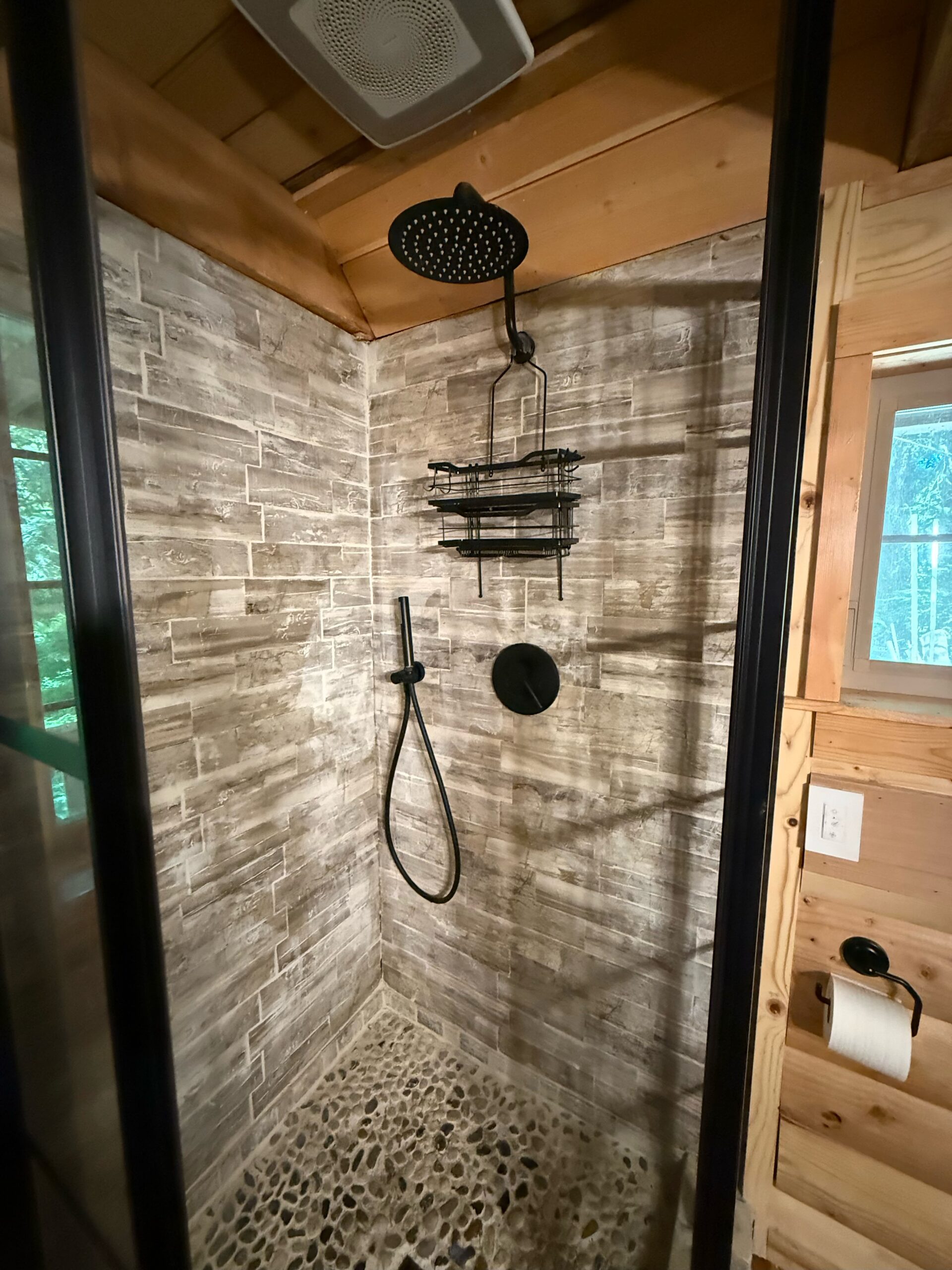 Modern shower with a rainfall showerhead, handheld sprayer, wall-mounted storage rack, stone floor, and wood and tile walls. A window and light switch are visible outside the shower.